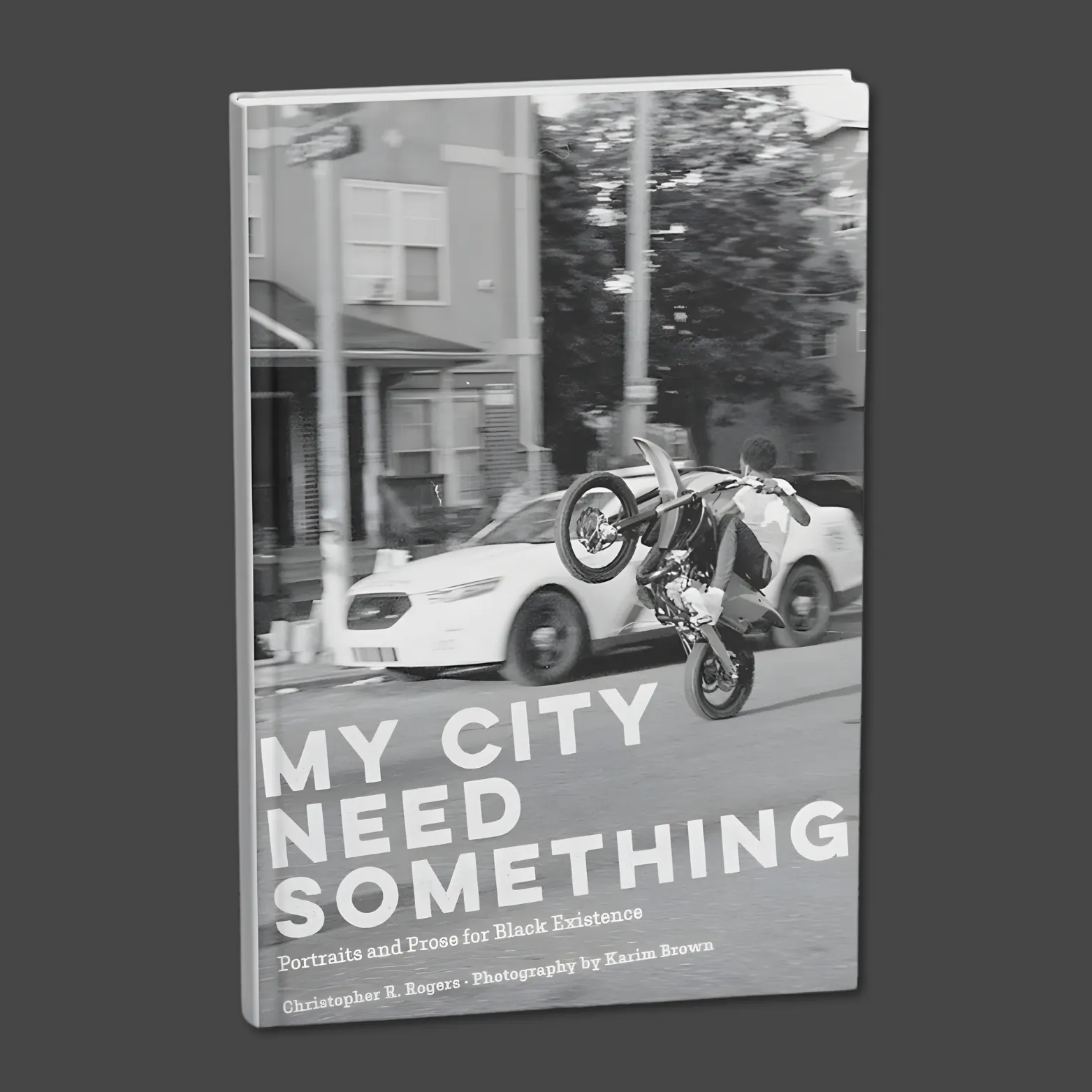 Black-and-white book cover showing a person doing a motorcycle wheelie on a city street, with the title “My City Need Something” and the subtitle “Portraits and Prose for Black Existence.”