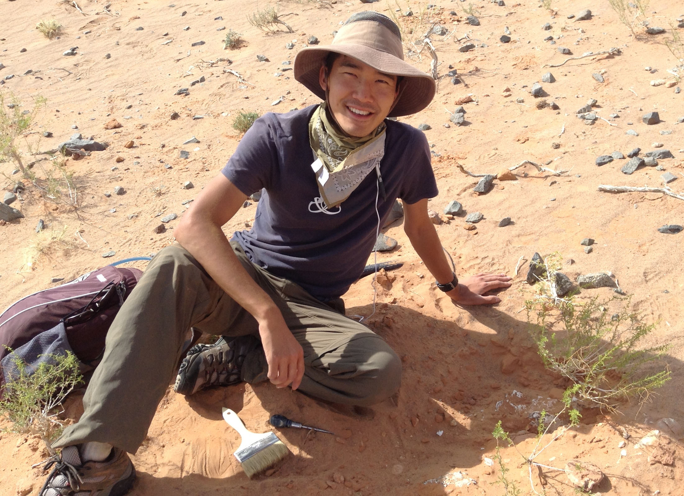 Smiling person wearing a wide-brimmed hat and outdoor field clothing, kneeling in a sandy desert landscape with a brush and tools nearby.
