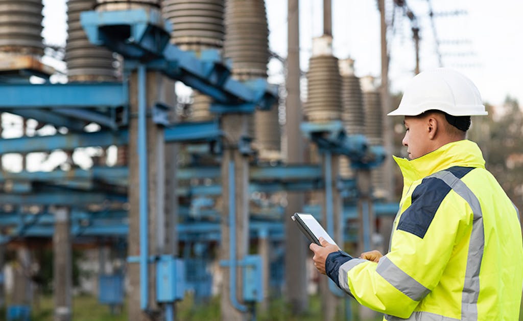 Electrical engineer wearing white hard hat and yellow safety vest inspects an electrical substation with blue components.