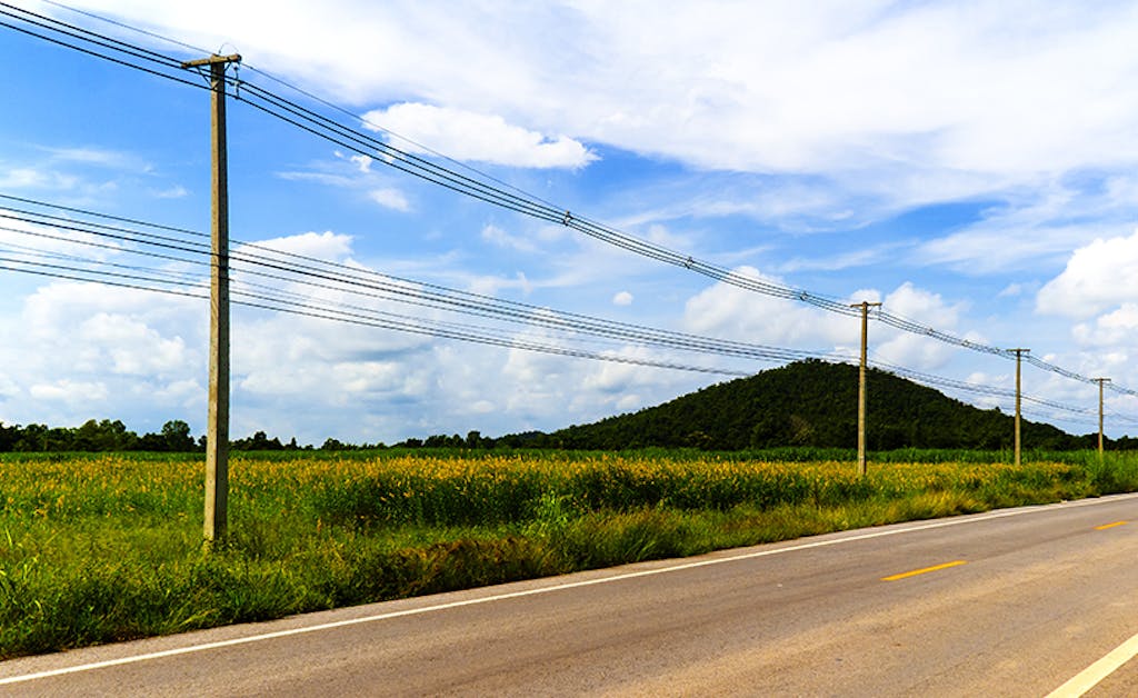 An electric pole stands tall along a serene rural road, bordered by lush green rice fields, under a clear blue sky with distant small mountains.