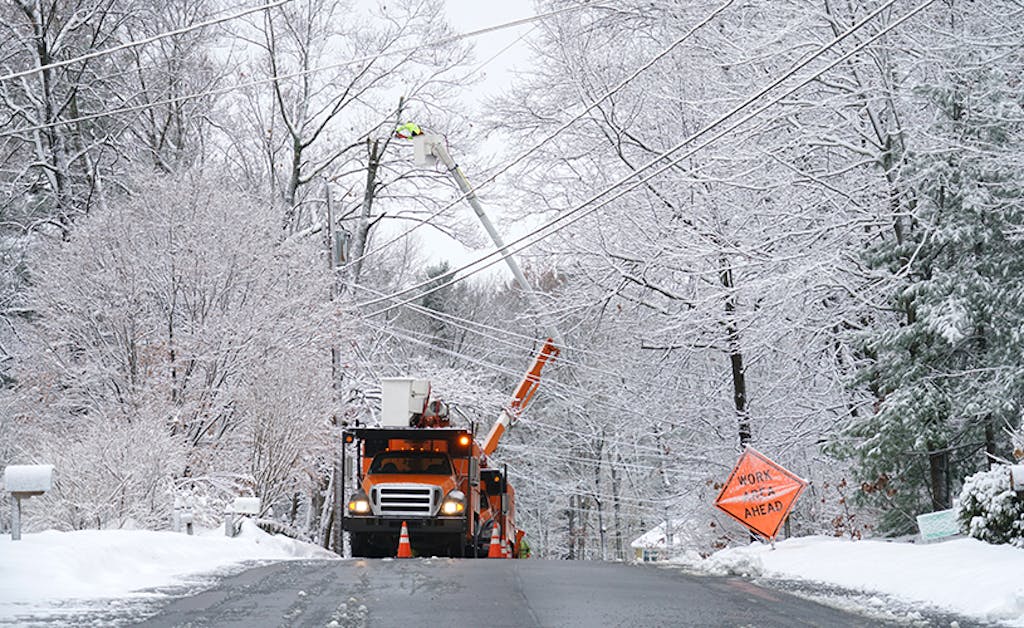 A bucket truck raises line workers to utility lines on snowy roads with large trees covered in snow.