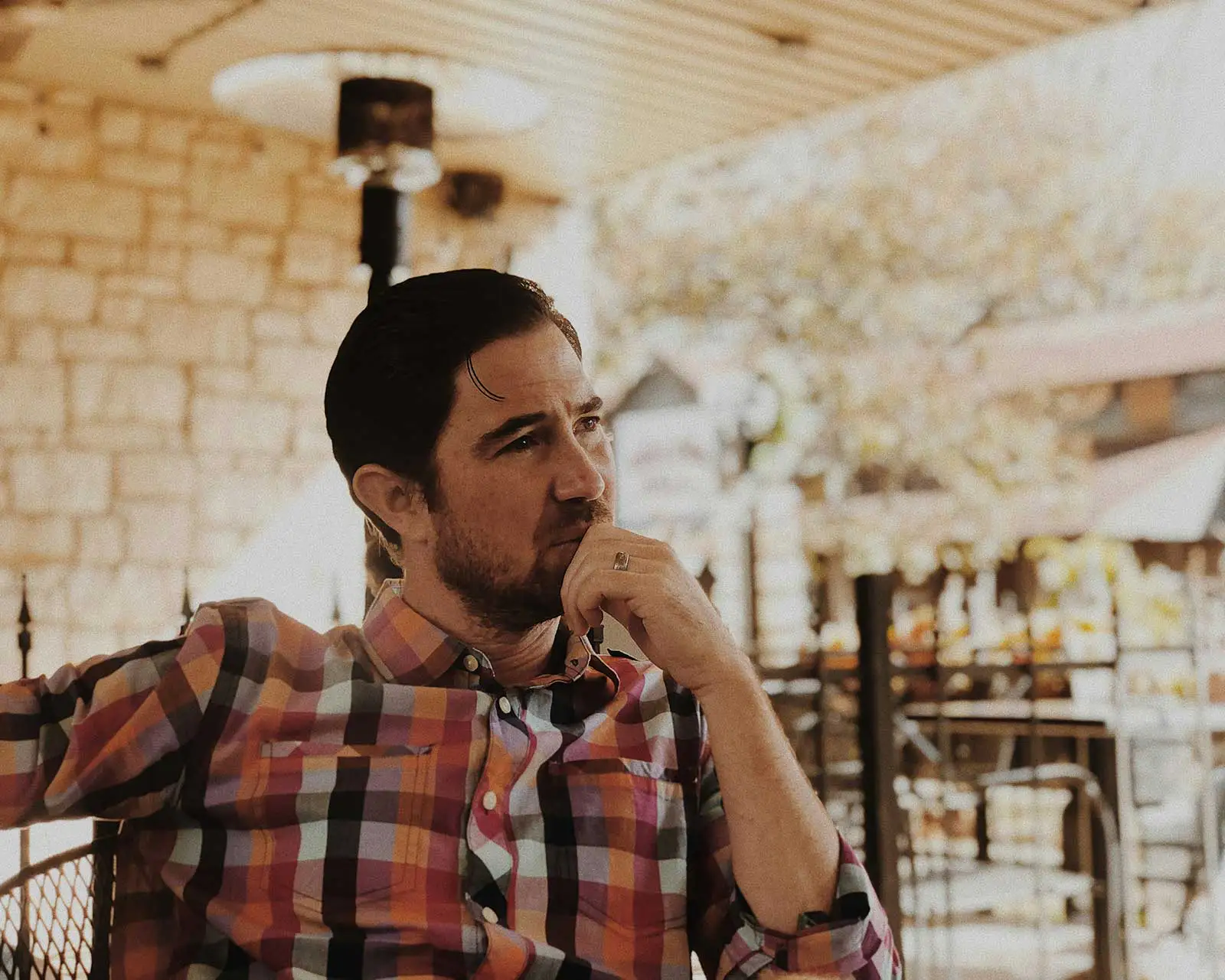 A man in a plaid shirt sits on a patio with his hand to his chin, deep in thought.