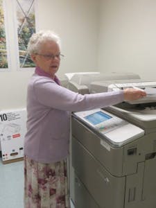 Sr. Angela copies information, prepared individual folders for a bulk mailing, and stacks the collated packets awaiting postage.