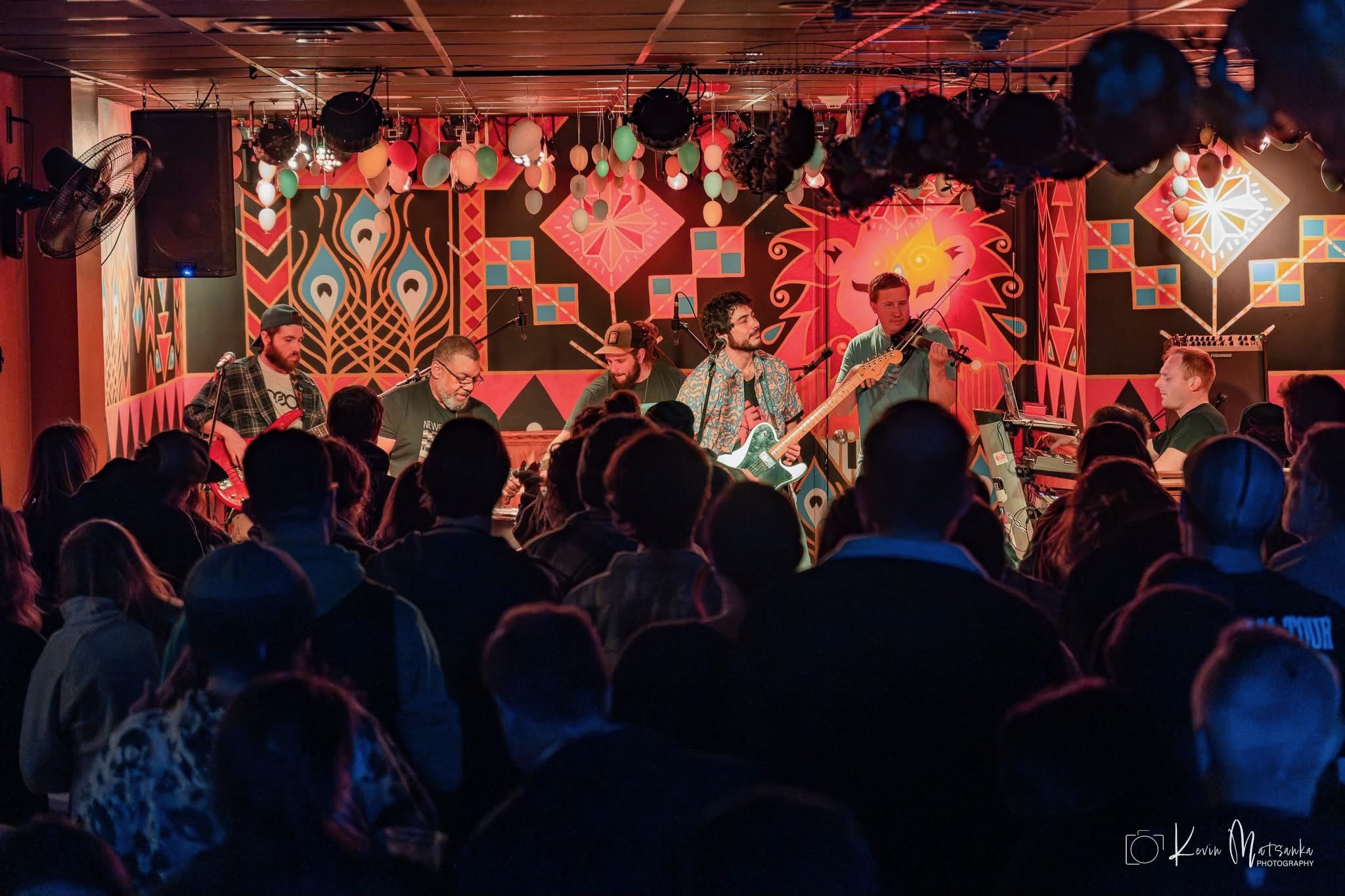 A band performing on stage in front of a geometric patterned backdrop, audience silhouetted in the foreground